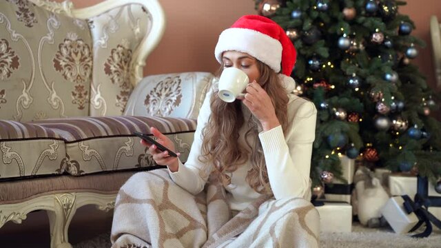 Christmas Woman dressed white sweater Santa hat sitting on floor near Christmas tree wrapped herself blanket Caucasian female relaxing winter holiday plaid drink hot coffee using smartphone