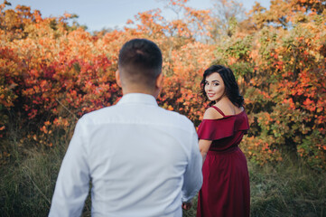 A beautiful girl in a red dress looked back at her beloved boyfriend, walking by the hands through the forest in autumn against the backdrop of nature.