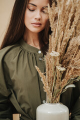 Close up portrait of a young woman with closed eyes holding cereal bouquet. She has long straight hair, wearing olive dress.