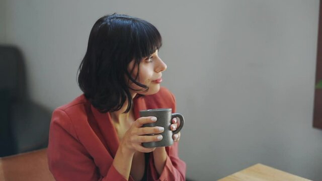Caucasian lady model holding a dark grey coffee cup looking at window.