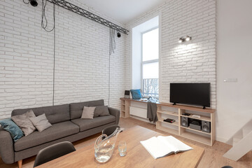 Small and stylish living room with white brick on the walls, gray sofa, big window and wooden dining table