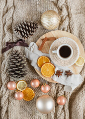 flat lay of a cup of coffee on wooden round tray on knitted beige blanket background with Christmas decorations around it: Christmas ornaments, pine cones, ribbons, dried oranges, cinnamon and anise