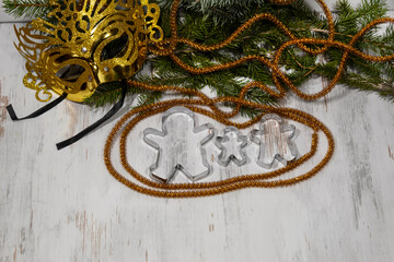 Fir branches and a carnival golden mask lie on a light background. In front, framed by tinsel, there are three metal forms in the form of gingerbread men.