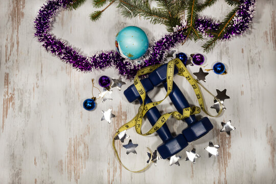 View from above. There is a centimeter on two blue dumbbells. Confetti in the form of silvery stars lies around. Nearby are spruce branches, New Year's tinsel and balls.