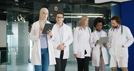 Mixed-races male and female healthcare professionals in medical coats discussing coronavirus treatment statistics while typing on tablet walking in clinic at workplace. Hospital workers concept - Powered by Adobe