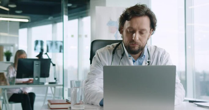 Portrait of handsome adult busy Caucasian male doctor sitting at desk in hospital office working and typing on laptop at workplace. Healthcare worker tapping on computer. Job concept