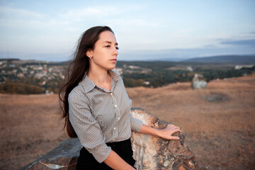 A young very beautiful girl is sitting on a large rock against the background of an autumn landscape and looks thoughtfully into the distance.