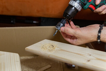  a man drills a big hole in the board on a cardboard box at home, DIY
