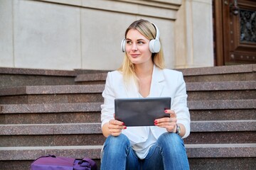 Young woman in headphones with digital tablet relaxing sitting on steps in city