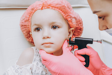 Charming little girl having an ear piercing process with special equipment in a beauty center by a...