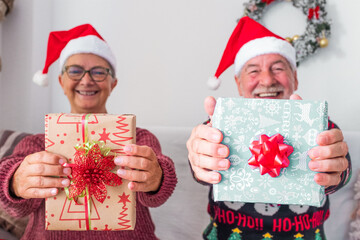 two mature people or seniors at home sitting on the sofa the christmas day opening and wrapping their presents or gifts together - cheerful and smiling people having fun