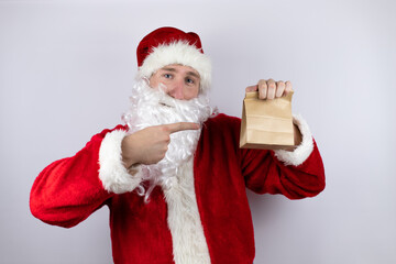 Man dressed as Santa Claus standing over isolated white background holding a paper bag and pointing the bag