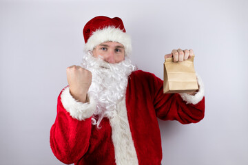 Man dressed as Santa Claus standing over isolated white background holding a paper bag very happy and excited making winner gesture with raised arms, smiling and screaming for success