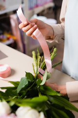 Obraz na płótnie Canvas Close up view of florist hand holding decorative ribbon, while tying stalks of bouquet in flower shop on blurred background