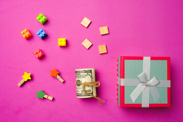 A flatlay picture of gift box with fake money, start clip, wooden tiles and building block on pink...