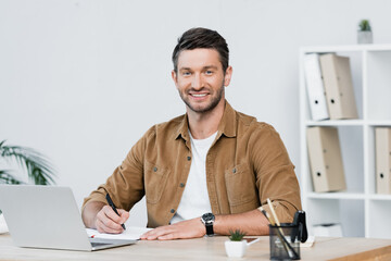 Fototapeta premium Smiling businessman looking at camera while writing in notebook at workplace with laptop on blurred background