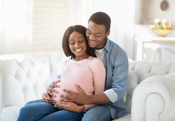 Smiling future mom and dad expecting baby, sitting on sofa at home and hugging