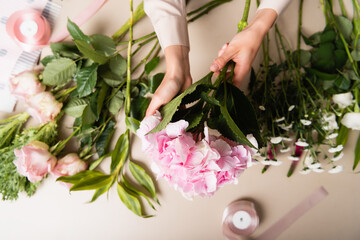 Cropped view of florist holding blooming hydrangea, while composing bouquets near flowers and decorative ribbons on desk