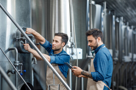 Millennial guy worker in apron turns a valve on big boiler for lager, manager with tablet controls process in interior
