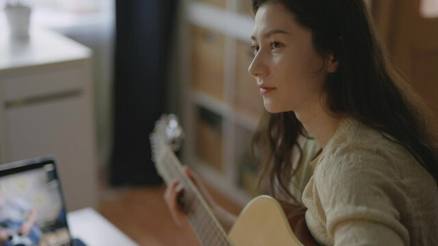 Young asian woman playing guitar at home. Aspiring musician learning to play musical instrument using laptop. Stay home, solo activity Pretty girl practising music with friend, online communication.