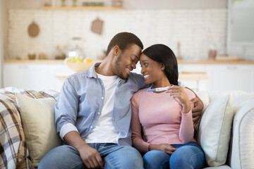 Loving African American couple with positive pregnancy test cuddling on sofa at home