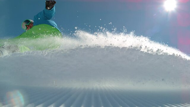 SLOW MOTION TIME WARP, LOW ANGLE, CLOSE UP, DOF: Athletic male tourist shredding the slopes sprays snow at the camera. Snowboarder carves along a groomed ski slope of a ski resort in the sunny Alps.