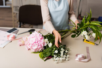 Cropped view of florist with roses taking chrysanthemums from desk with tools near decorative ribbons on blurred background