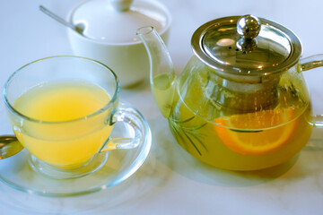 winter citrus healthy tea with rosemary, lemon and orange, in a glass teapot and a cup. beautiful tea-set table, lemonade yellow, hard light, copy space, selective focus