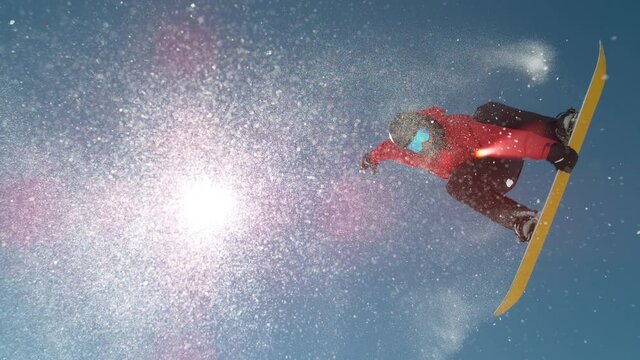 SLOW MOTION TIME WARP, LENS FLARE, BOTTOM UP: Snowflakes sparkle in the sunshine as male rider does snowboarding tricks. Fearless snowboarder does a spin trick while riding off a big air kicker.