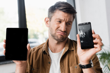 Sad businessman holding smartphone with blank screen while looking at smashed cellphone on blurred background