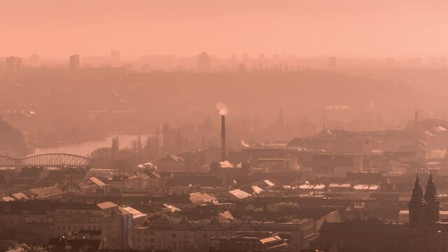 The orange haze hanging above Prague obscuring the view. Smoke rising above the rooftops and from the chimneys. Silhouettes of high-rise buildings seen in the background.