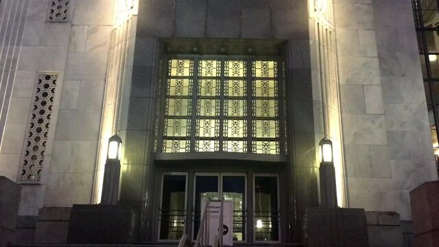 Grand entrance to historic art deco building, illuminated at night.  Art deco ironwork grill with interior light behind, doorway flanked by marble columns.  Zoom out, 4 K.