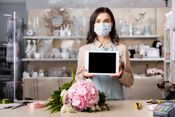 Florist in medical mask showing digital tablet near table with bouquet, tools and decorative ribbons on blurred background