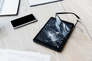 Eyeglasses on smashed digital tablet near documents and digital devices on wooden background