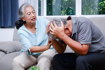 A sick Asian elderly man put his hand on his head. He is sitting on the sofa in the house. The wife is next door to take care. Concept of health care in retirement age