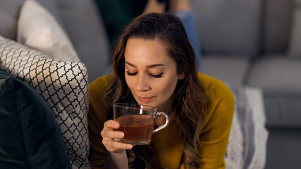 Young pretty woman drinks a cup of tea while lying on the couch - home shooting