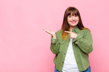 overweight woman smiling cheerfully and pointing to copy space on palm on the side, showing or...