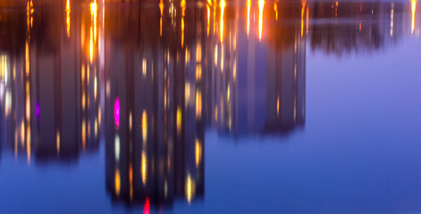 Silhouettes of multi-storey houses with bright light in the windows reflect on the blue water surface in the lake. Big city in details