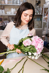 Smiling brunette florist looking at bouquet near desk with stalks on blurred racks on background