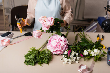 Cropped view of florist holding roses and secateurs near desk with flowers, decorative ribbons and tools on blurred background