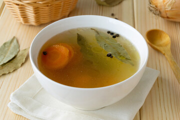 Meat bone broth with vegetables and spices in white bowl on wooden background
