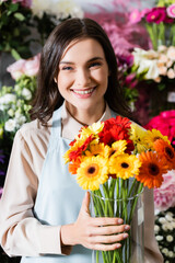Smiling female florist looking at camera while holding vase with gerberas with blurred range of flowers on background