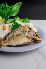 Fried two short bodied mackerel fishes and vegetable on white plate with black and white background.