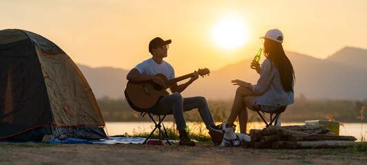 Asian couple with dog playing guitar and drinking beer beside their tent campsite panorama