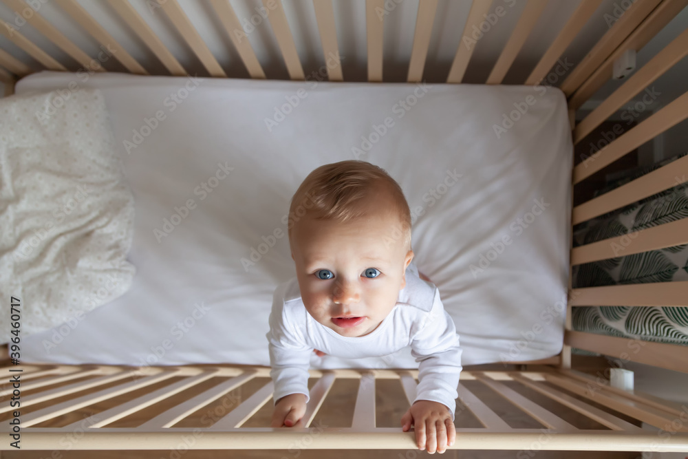 Wall mural blond cute llittle kid in white bodysuit stands in wooden bed