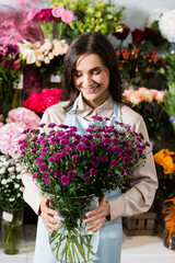 Front view of happy female florist looking at purple chrysanthemums in vase near blurred racks of flowers on background