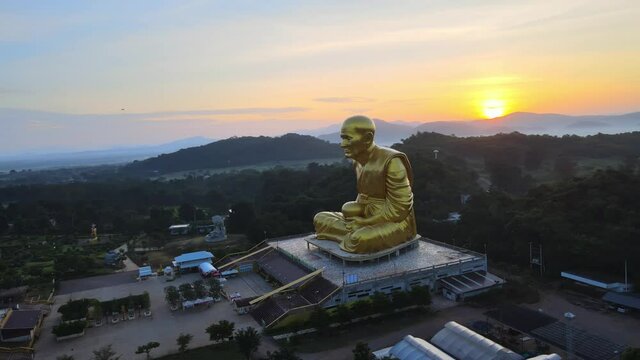 4k Aerial drone footage of a huge monk statue surrounded by mountains of Khao Yai at dusk in Thailand. Panning left to right around Luang Pu Thuat at dawn. Image of man made of gold. Medium shot