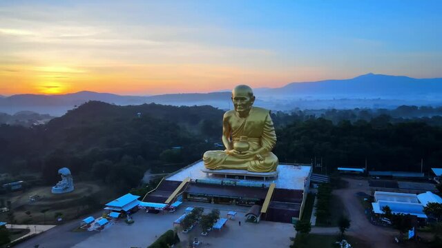 4k Wide Aerial shot of a Big Luang Por Tuad monk statue surrounded by mountains of Khao Yai at dusk in Thailand. Panning around of Luang Pu Thuat at dawn. Image of buddha disciple made of gold.