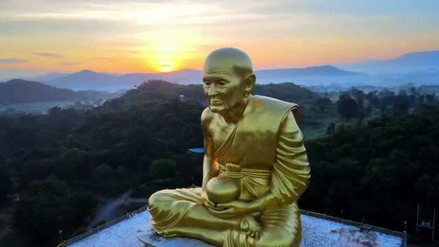 4k Aerial Luang Pu Thuat image at dawn. Drone footage of a huge golden monk statue surrounded by mountains of Khao Yai at dusk in Thailand. Panning around of the famous buddha disciple made of gold