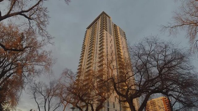 Stationary camera pan out upwards of clouds moving steady around a modern futuristic luxury apartment building next to vintage towers as sun sparkles light to the center surrounding by bare fall trees